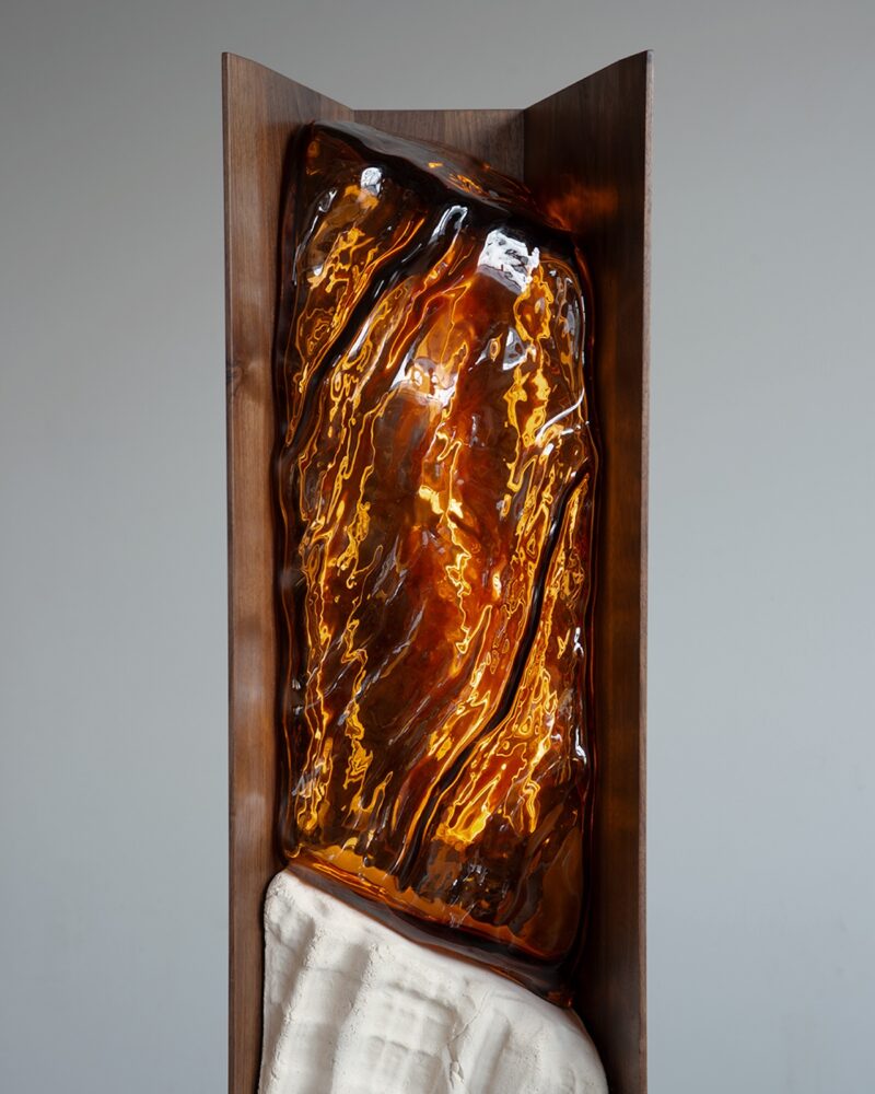 A vertical sculpture featuring a large, amber-colored, translucent resin form with flowing textures set in a brown frame, above a white, textured base.