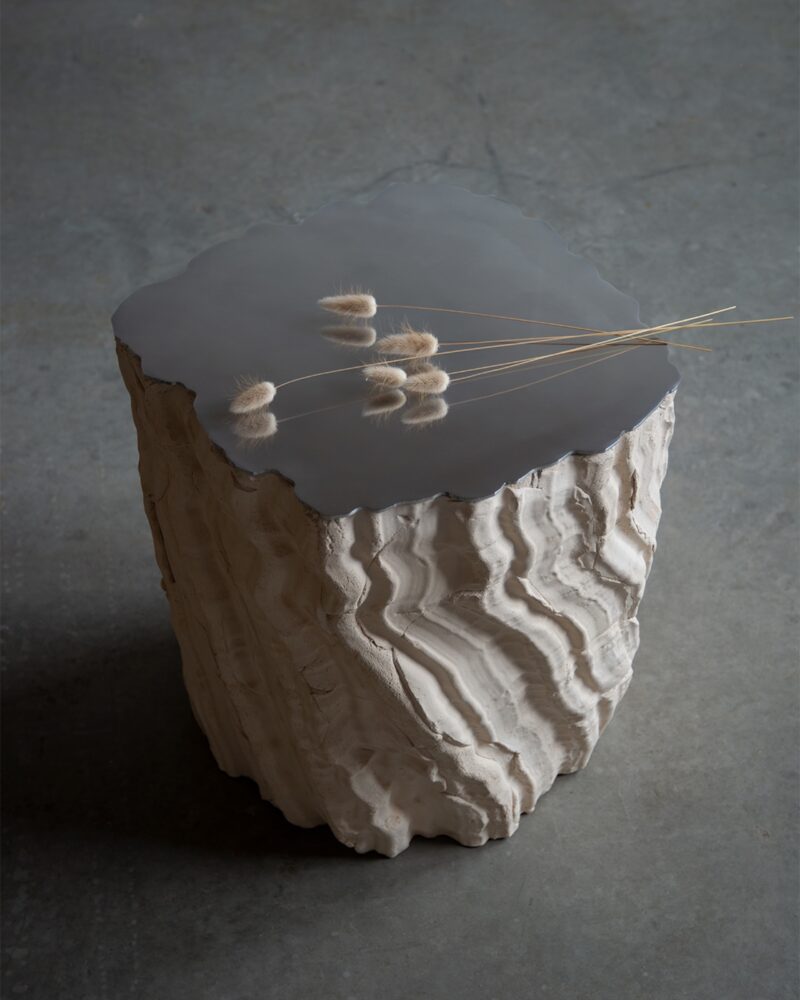 A textured, sculpted side table with a smooth gray top and rough, wavy white sides, holding a few dried plants, sits on a concrete floor.