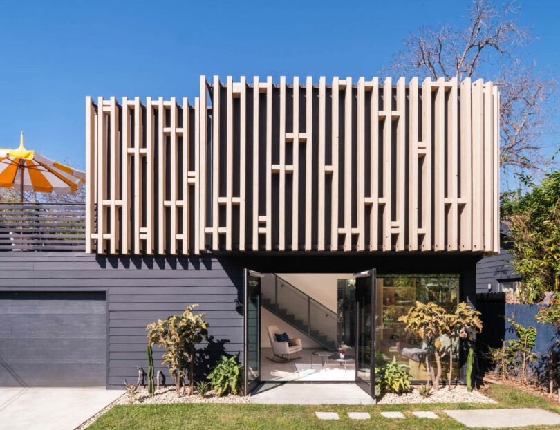 Modern two-story house with vertical wooden slats on the upper facade, large glass doors on the lower level, and a small garden in front.
