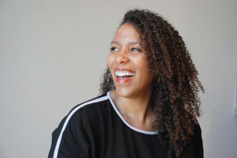 A woman with curly hair wearing a black top with white trim is smiling and looking to the side against a plain light background.