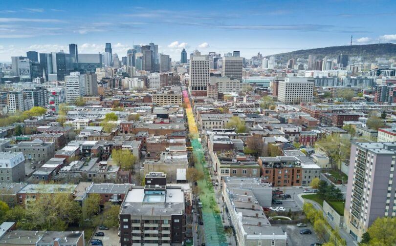 Aerial view of a cityscape with a long, colorful rainbow-painted street running through residential and commercial buildings, with skyscrapers and hills in the background.