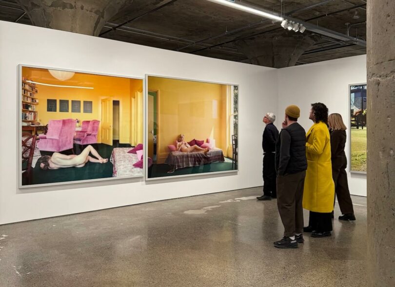 Four people view large, framed photographs of figures in bedrooms on display in a modern art gallery with concrete floors and exposed ceilings.