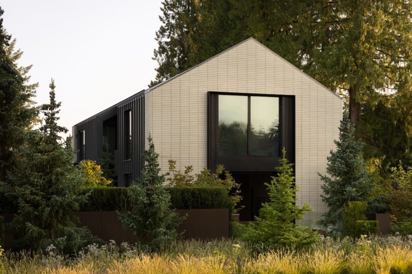 Modern two-story house with light-colored brick exterior, large windows, and dark accents, surrounded by trees and landscaped greenery.