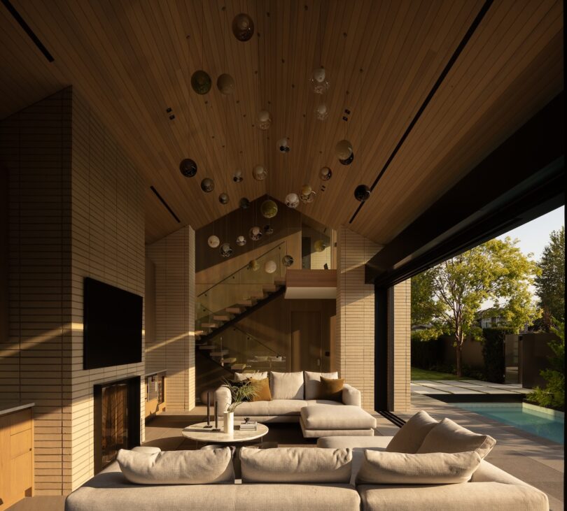 Modern living room with light wood ceiling and walls, sectional sofa, glass stair railing, wall-mounted TV, and open view to outdoor pool area and trees.