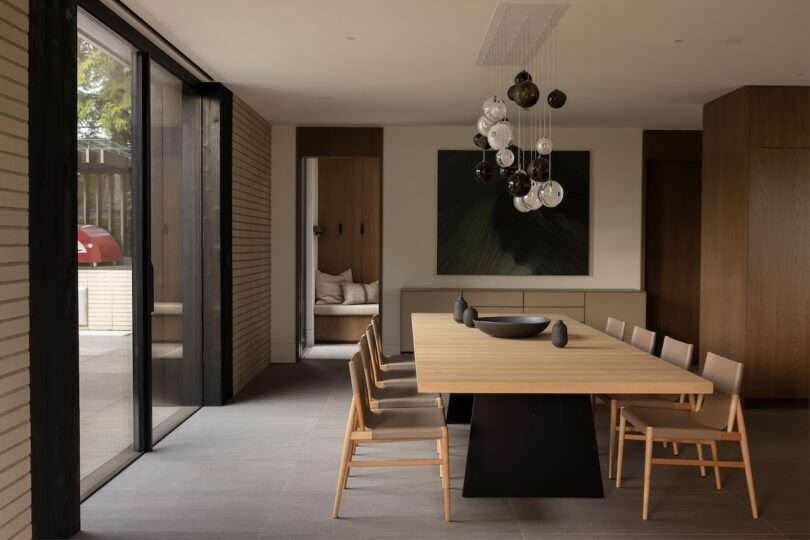 Modern dining room with a light wood table, six chairs, abstract art on the wall, and cluster pendant lights above. Large sliding glass doors open to an outdoor area.