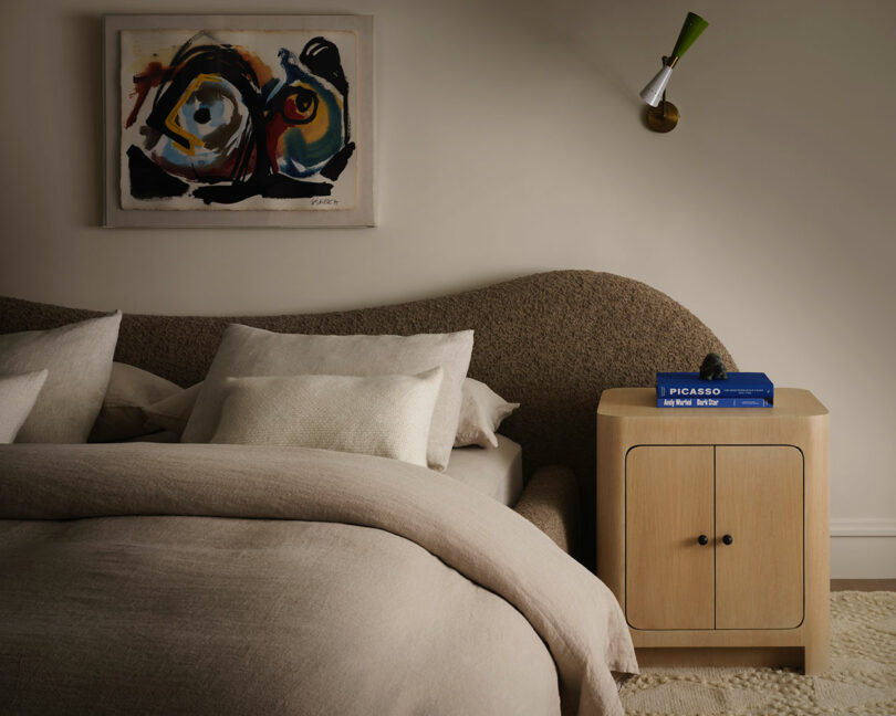A neatly made bed with neutral bedding, a curved textured OSKLO headboard, a wooden nightstand with books, and an abstract painting hanging above.