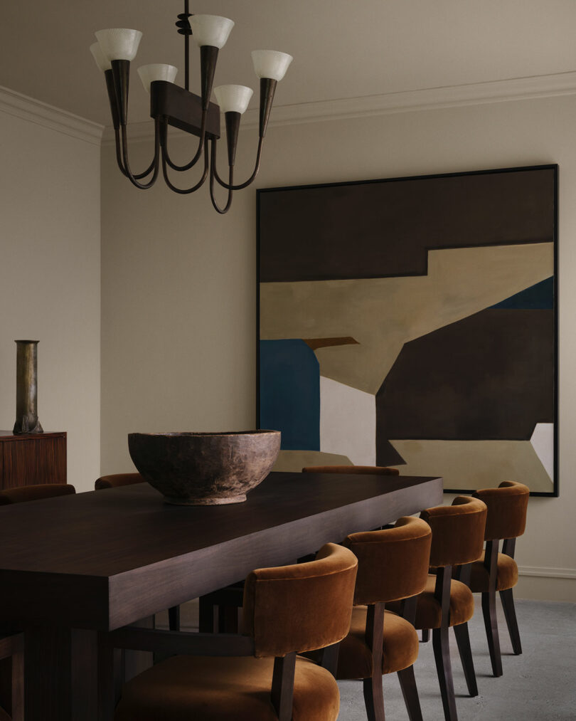 A modern OSKLO dining room with a dark wooden table, rust-colored chairs, a large abstract painting, and a striking black chandelier.
