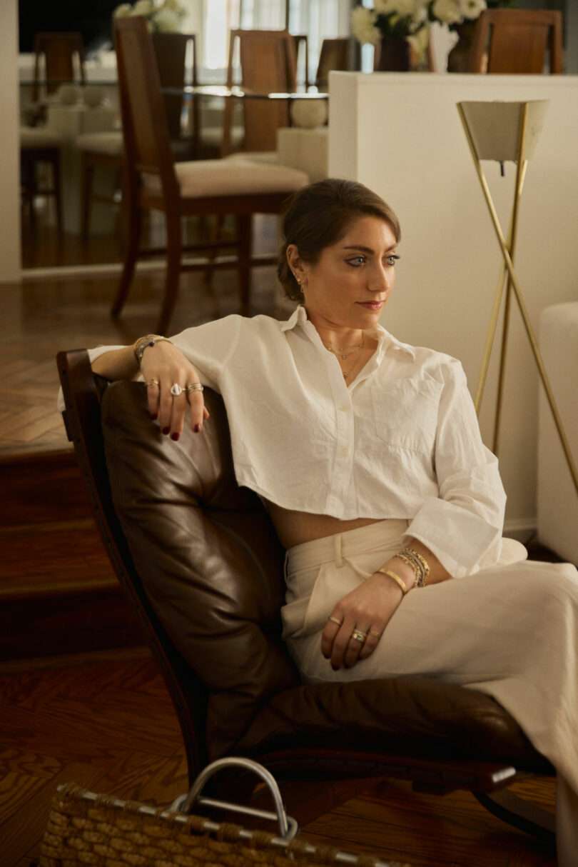 Coco Greenblum sits in a brown leather chair, wearing a white blouse and light pants, in a modern, well-lit living room with wooden floors and dining furniture in the background.