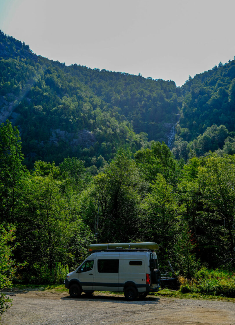 A white campervan parked on a dirt clearing surrounded by dense green trees, with forested mountains and a waterfall in the background—perfect for your next Friday Five adventure.