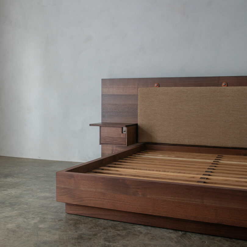 A Friday Five favorite: this wooden platform bed frame features a slatted base, built-in headboard, side shelf, and beige upholstered panel, all set against a plain wall and concrete floor.
