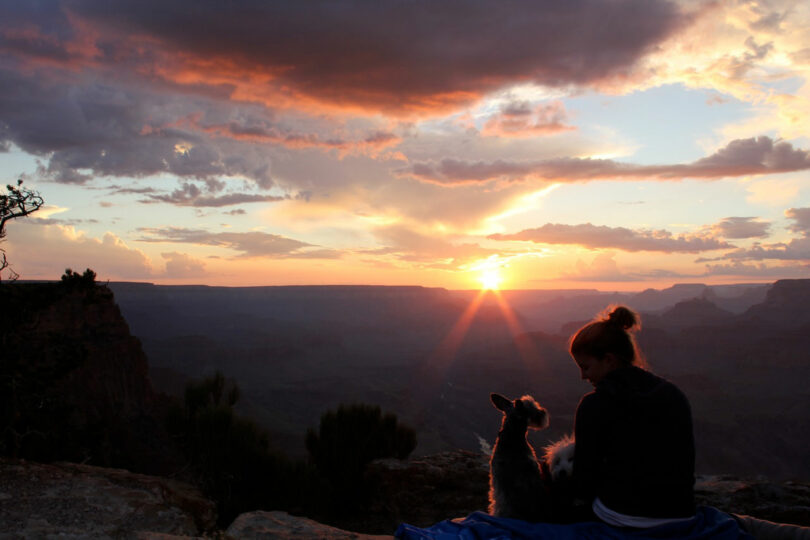 A person sits on a blanket with a dog, watching the sunset over a canyon landscape as clouds drift above distant cliffs—a perfect Friday 5 moment to unwind and soak in nature’s beauty.