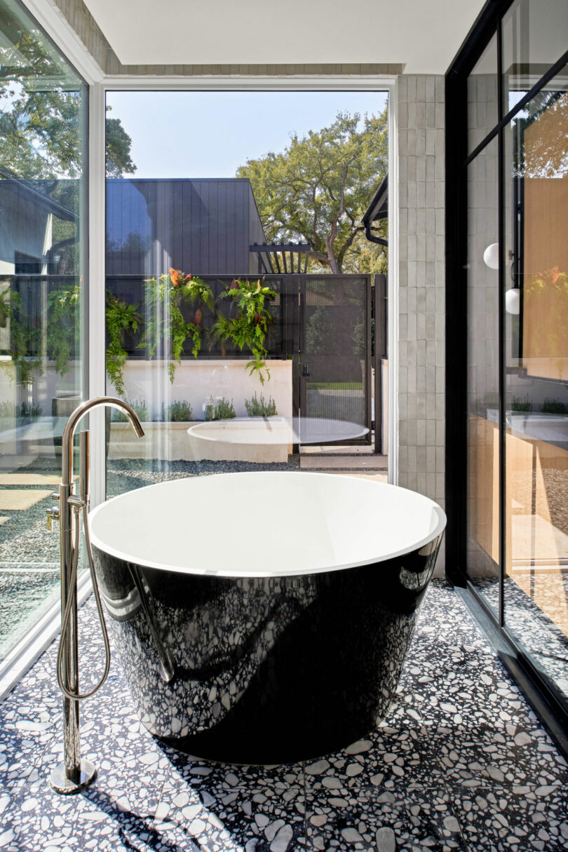 A modern bathroom with a black and white freestanding bathtub next to a floor-mounted faucet, large windows, and outdoor garden views—perfect inspiration for your Friday 5 mood board.