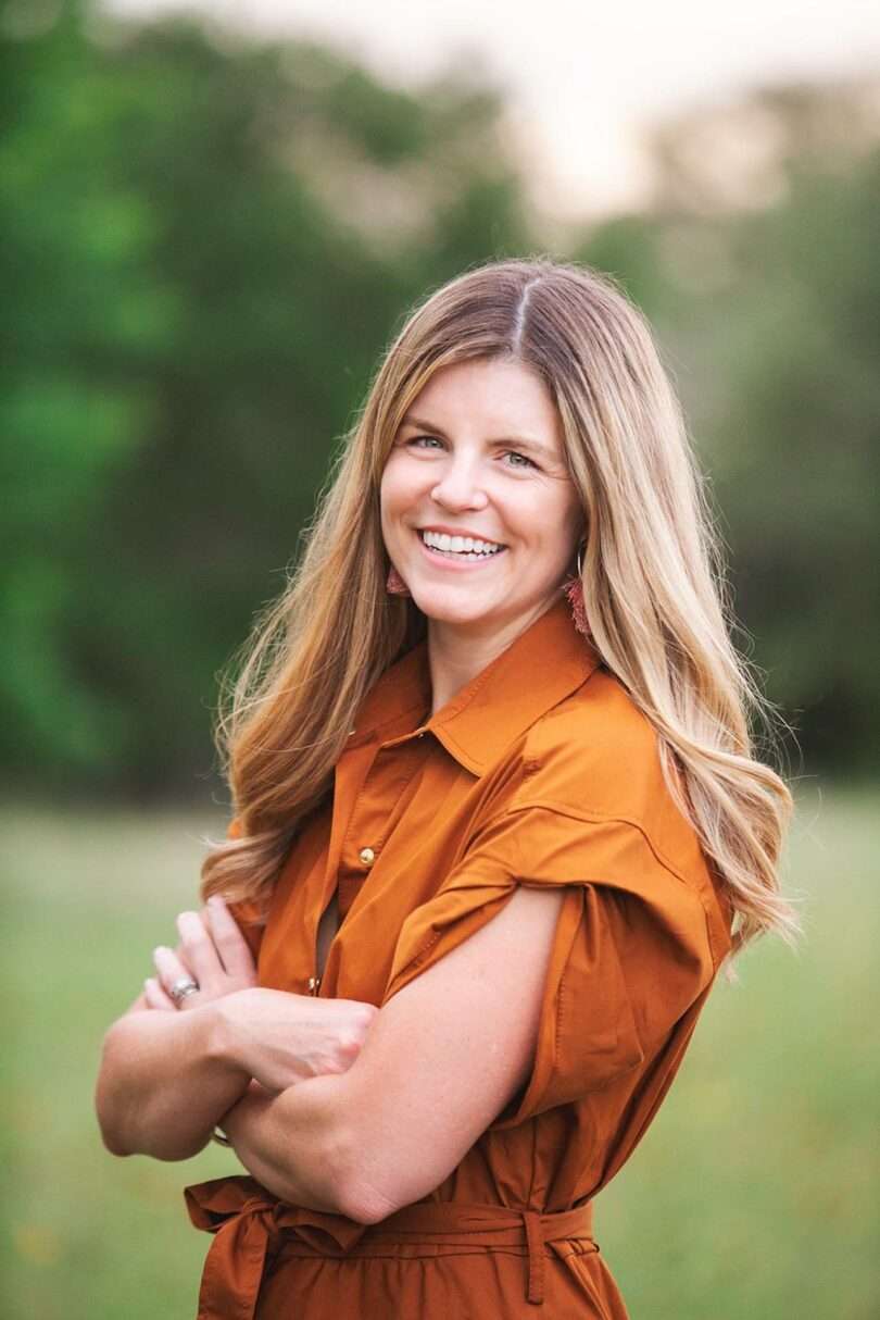 A woman with long blonde hair, wearing an orange dress, stands outdoors with arms crossed and smiles at the camera. Green blurry trees are in the background, capturing a relaxed Friday 5 vibe.