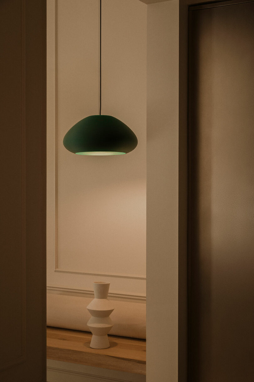A green pendant lamp hangs above a wooden shelf holding a white geometric vase in a softly lit, minimalist interior space.