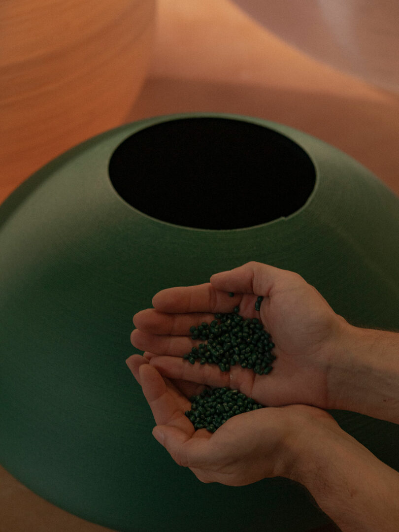 A person holds small green pellets in their cupped hands above a large green container with a round opening.