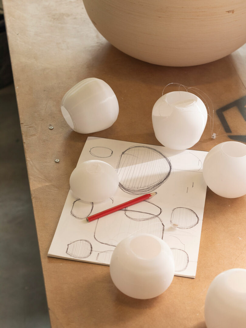Several white round objects are placed on a table next to a sheet of paper with sketches of similar shapes and a red pencil.