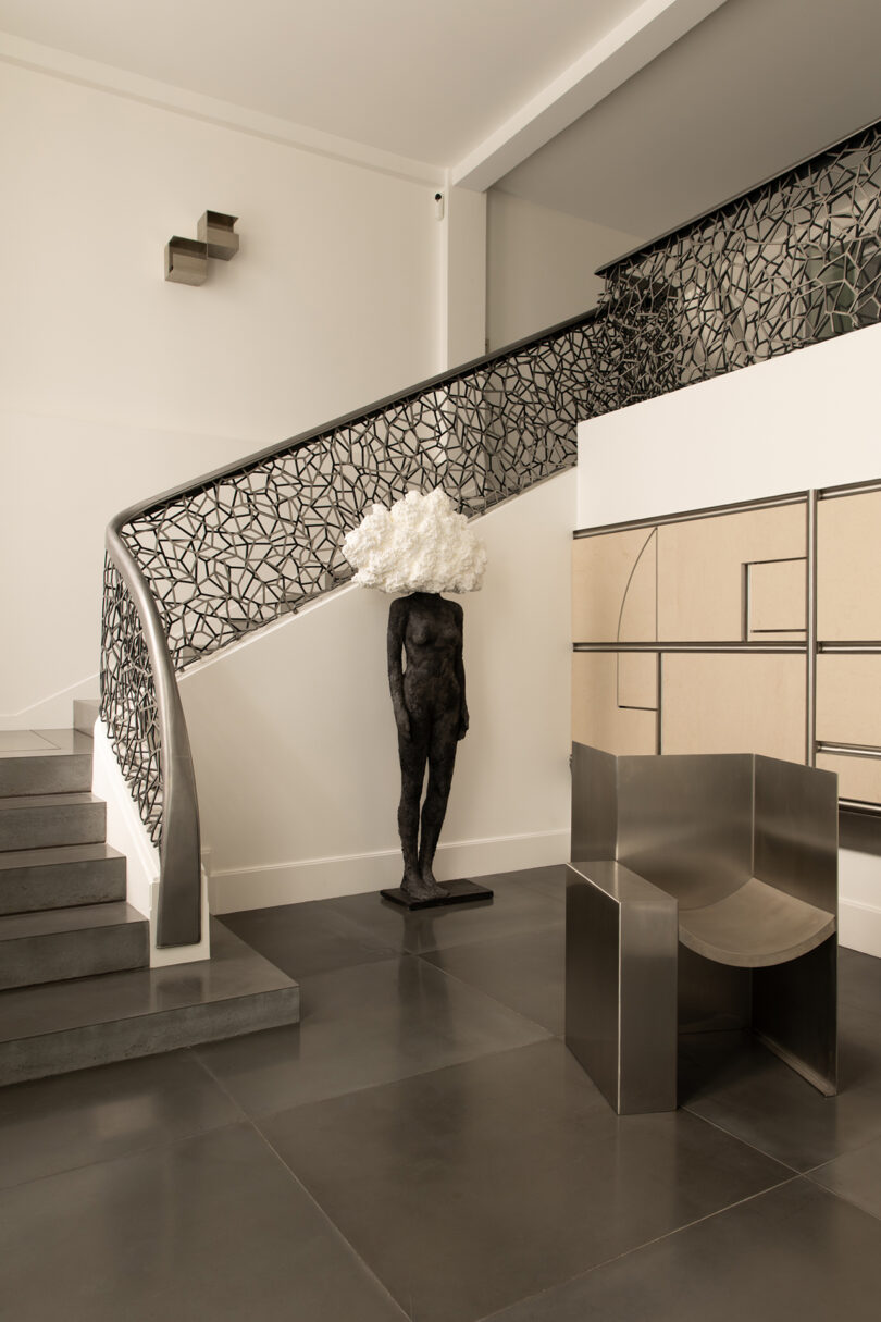 Modern interior with a sculptural staircase, geometric wall art, a black statue with a white cloud-like head, and a minimalist metallic chair on a tiled floor.