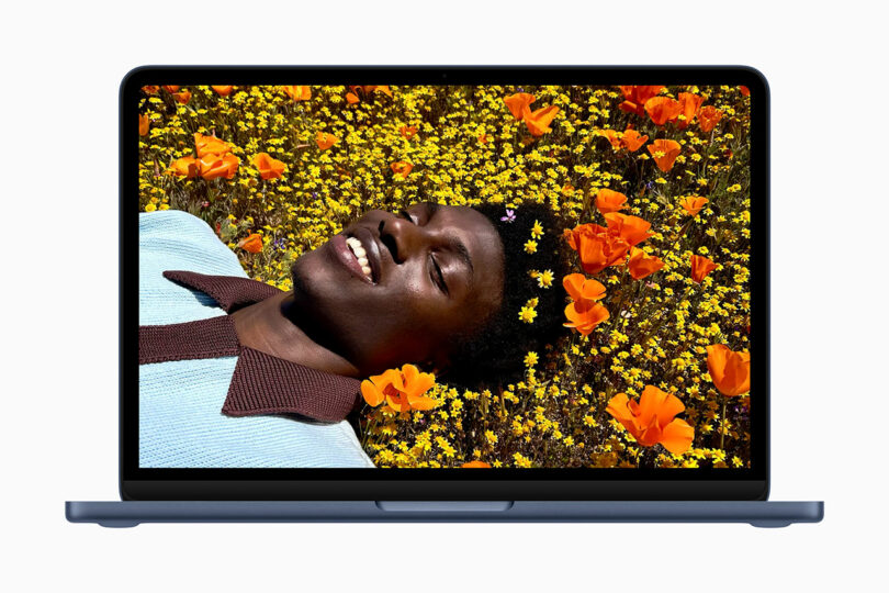 A laptop, possibly an Apple device, displays an image of a person lying in a field of yellow and orange flowers, smiling and looking upward.