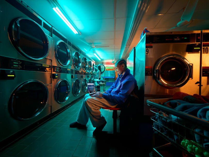 A person sits on a bench in a laundromat at night, using an Apple laptop. Large washers and dryers line the walls under blue-green fluorescent lighting.