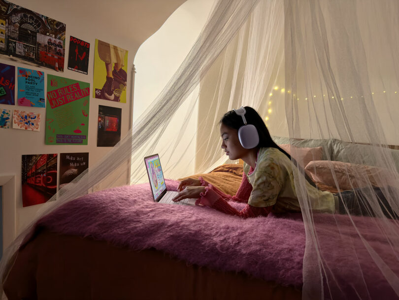 A girl wearing headphones uses an Apple laptop while lying on a bed with a pink blanket, surrounded by posters and string lights, under a sheer canopy.