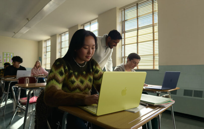 A student uses a yellow Apple laptop at her desk while a teacher assists another student in a classroom with several other students working.