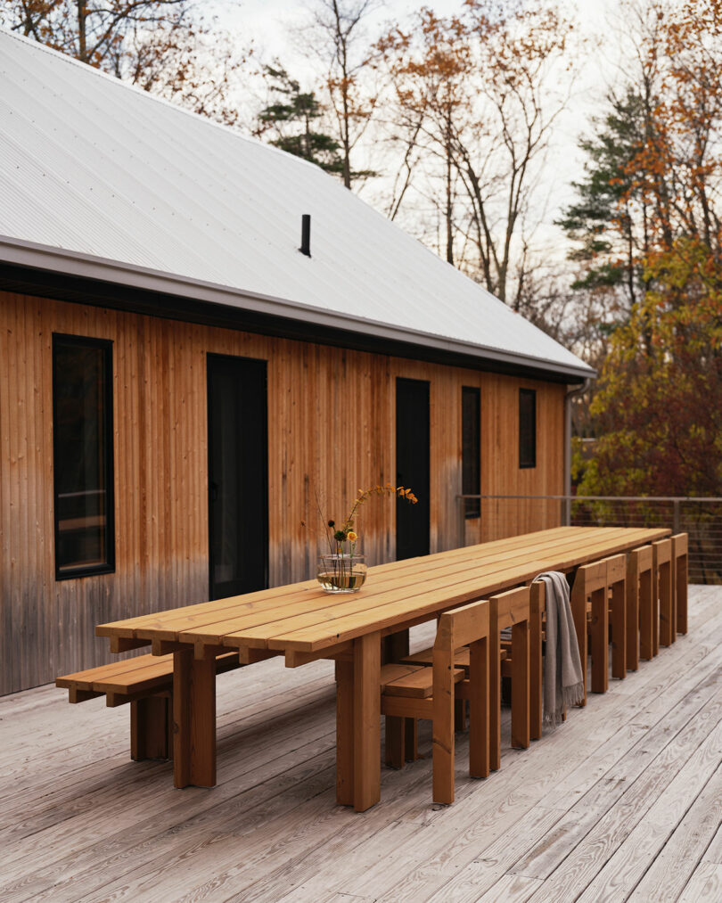 A long wooden Design Within Reach table with benches sits on a deck beside a modern wood-paneled building; a vase with flowers and a draped blanket are on the table. Trees with autumn leaves are in the background.