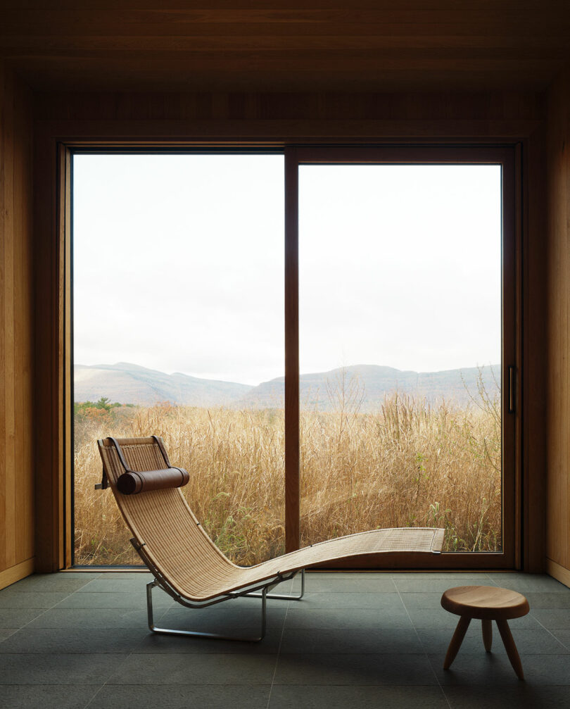 A minimalist room with a Design Within Reach wicker chaise lounge and a small wooden stool near a large window overlooking dry grass and distant mountains.