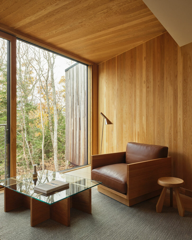 A modern room with Design Within Reach wood-paneled walls, a large glass window, a brown leather armchair, a glass coffee table, a wooden stool, and a floor lamp overlooking trees outside.
