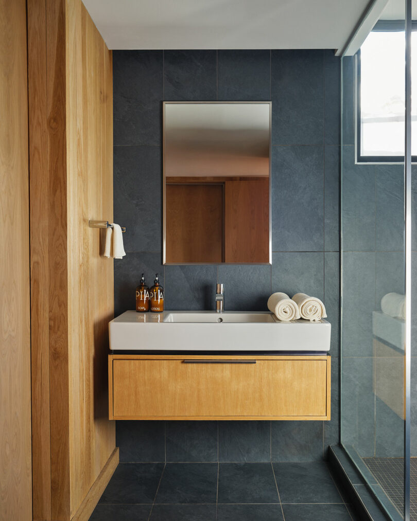 Modern bathroom with a rectangular mirror above a white sink and wooden vanity, dark tile walls, soap dispensers, rolled towels, and a Design Within Reach-inspired glass shower enclosure.
