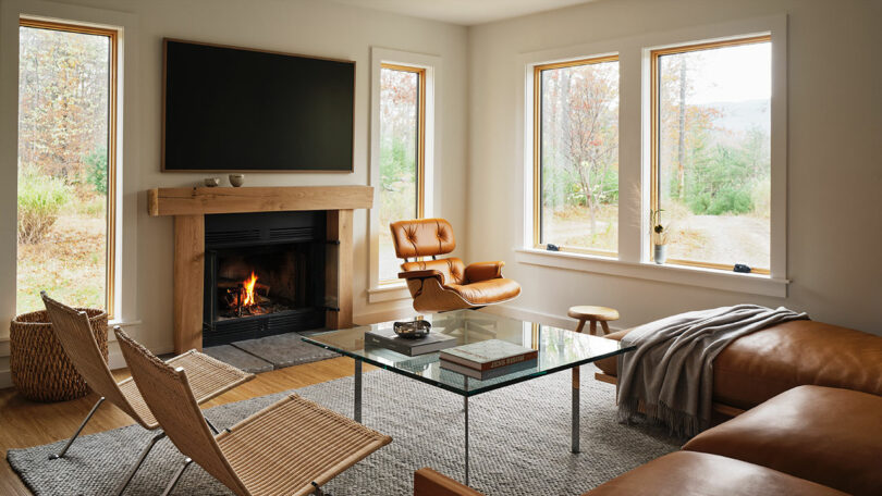 Modern living room with a fireplace, leather furniture from Design Within Reach, glass coffee table, and large windows revealing trees and an outdoor view.