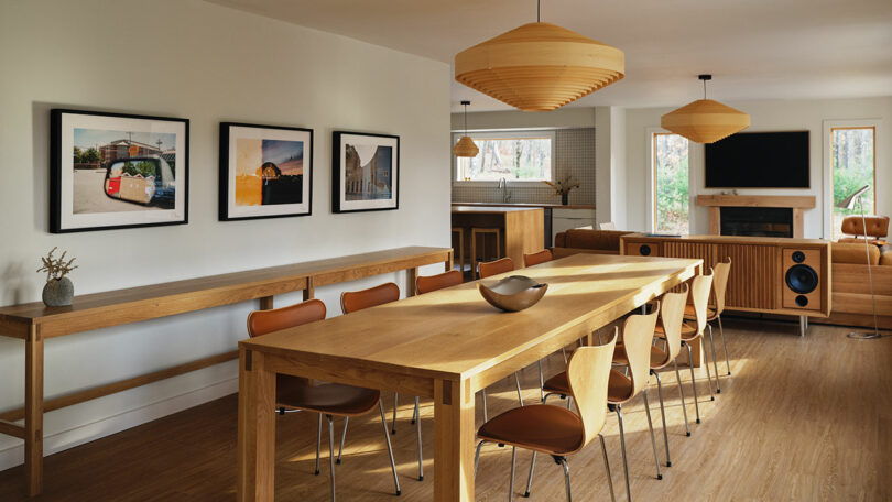 A modern dining and living area featuring a long wooden table, brown chairs, wood furniture from Design Within Reach, pendant lights, wall art, and a fireplace with a TV above it.