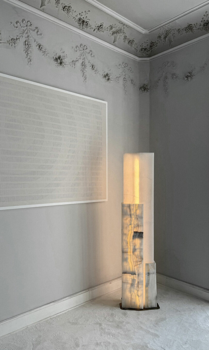 A modern ATRA floor lamp with stacked translucent stone blocks emits soft light in a minimalist, ornate room with detailed ceiling molding and monochromatic wall art.