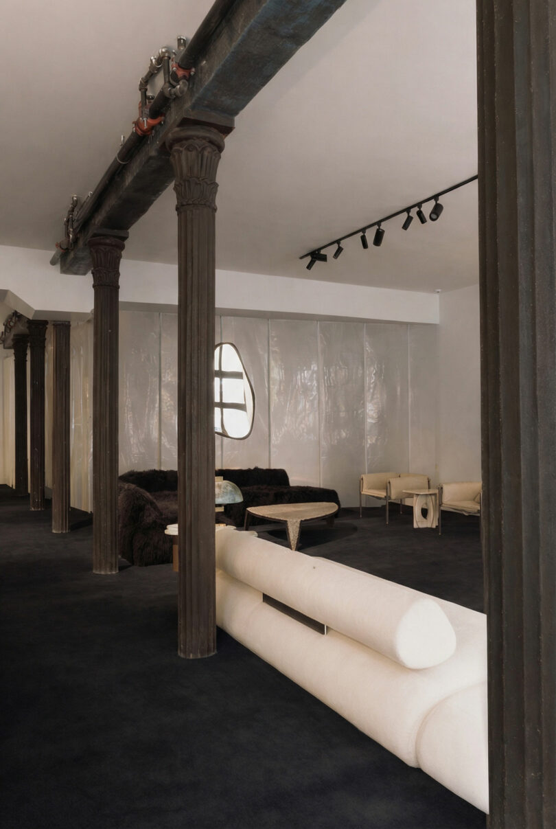 Modern lounge with dark columns, black carpet, ATRA cream and black sofas, a marble table, and a translucent wall with an oval window. Track lighting is mounted on the ceiling.