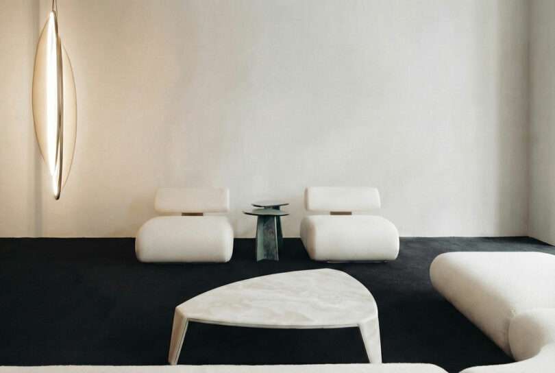 Minimalist living room with two modern white ATRA chairs, a small sculptural side table, a triangular coffee table, and a unique vertical light fixture against a plain white wall and dark floor.