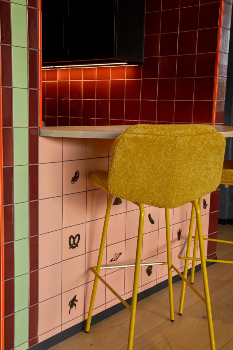 A yellow upholstered Powell barstool sits at a counter, with a tiled wall featuring red, peach, and green tiles, some decorated with black food illustrations.