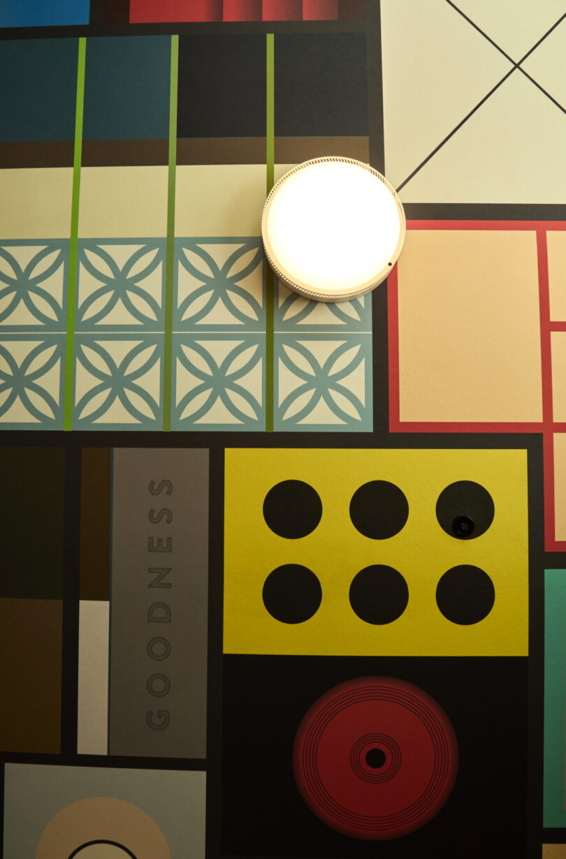 Geometric patterned wall with a variety of colored shapes and a lit round Powell wall sconce at the top center. The word “GOODNESS” is printed vertically on one section.