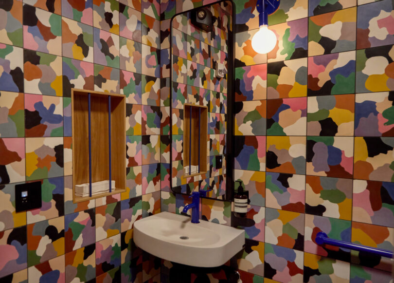 Bathroom with a small sink, mirror, and shelves, surrounded by walls covered in Powell’s multicolored, abstract-patterned tiles.