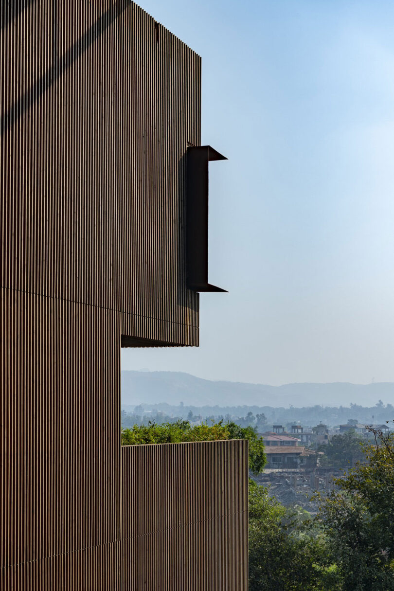 A modern building by Malik Architecture with vertical wooden slats is shown in the foreground, while trees and distant hills are visible beneath a clear blue sky in the background.