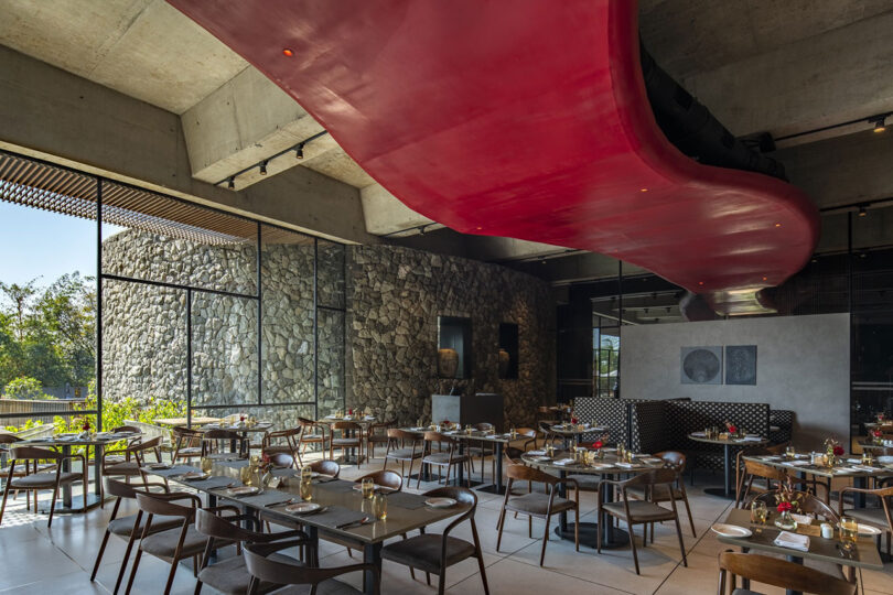 Modern restaurant interior by Malik Architecture, featuring stone walls, wooden tables and chairs, large windows, and a striking curved red ceiling. Tables are set for dining.