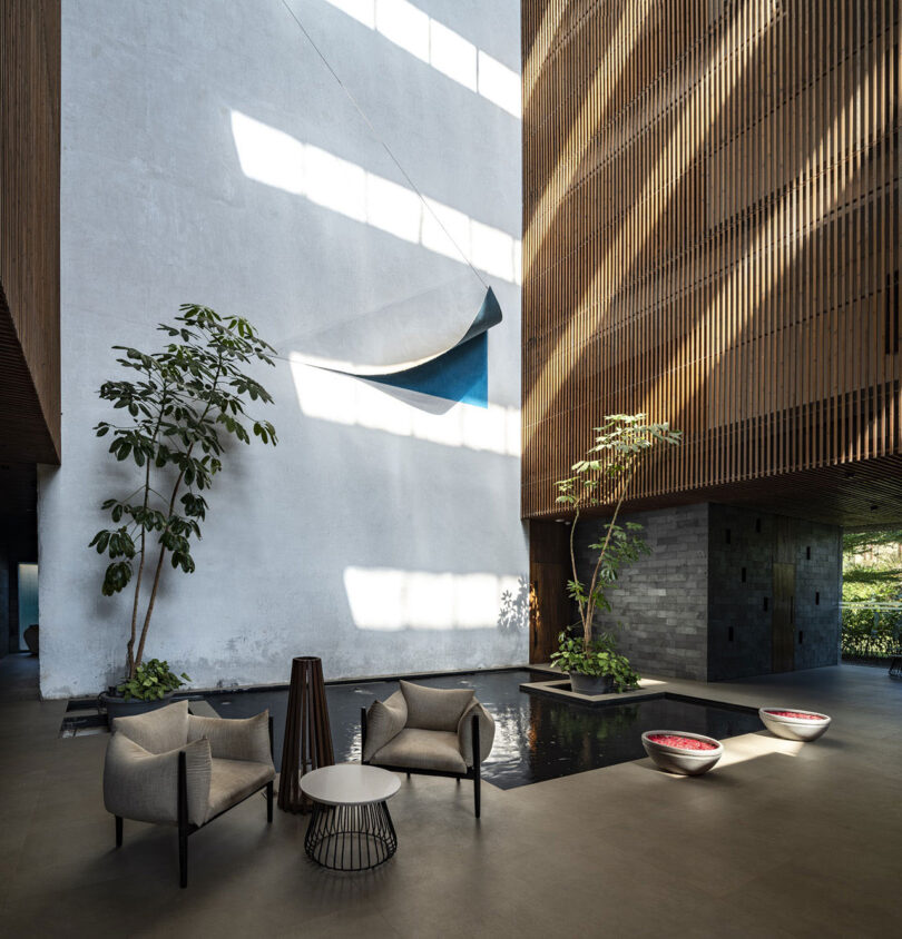 Modern lobby by Malik Architecture with two armchairs, a small table, indoor plants, a shallow reflecting pool, and large windows casting geometric shadows on the walls.