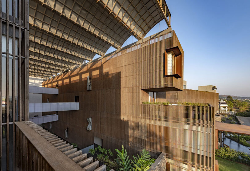 Modern building by Malik Architecture featuring a large, angled metal roof, vertical wooden slats on the façade, and extended window detail; clear sky and greenery visible in the background.