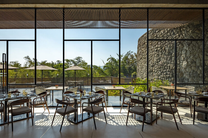 Modern restaurant interior by Malik Architecture featuring glass walls, wooden tables and chairs, plus an outdoor patio with umbrellas and greenery visible through expansive windows.