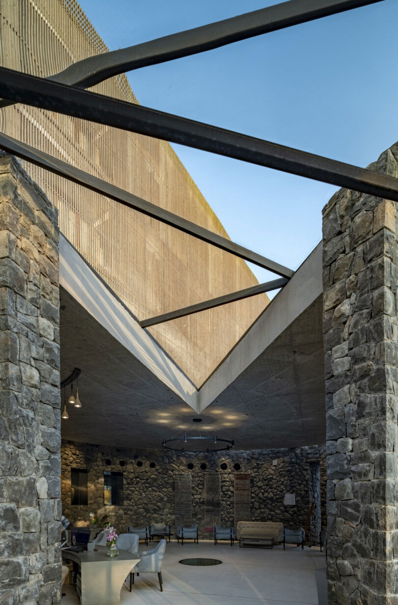 Outdoor seating area with stone walls and exposed steel beams, partially covered by a concrete roof under a clear blue sky, exemplifying the signature style of Malik Architecture.