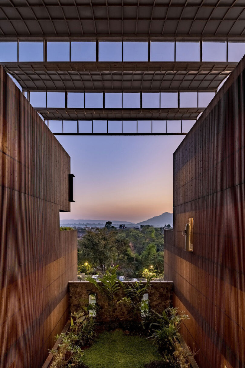 View between two modern wooden walls—Malik Architecture’s signature—overlooking greenery, distant hills, and a colorful sky at sunset, all elegantly framed by a metal roof structure above.