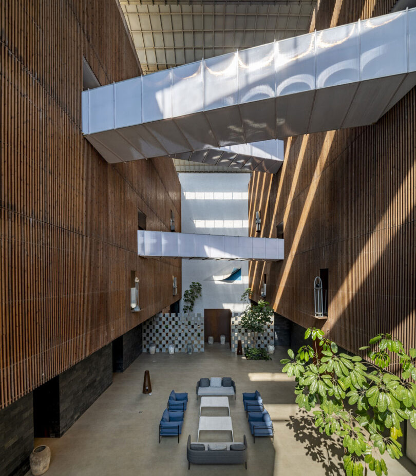 A modern atrium by Malik Architecture with wooden slat walls, overhead walkways, lounge seating, potted plants, and natural light streaming through windows and skylights.