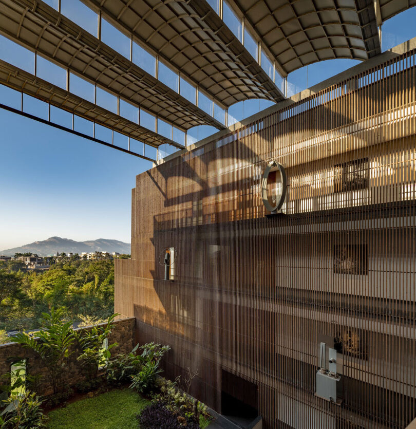 A modern building by Malik Architecture with a metal mesh façade and circular windows, partially covered by an overhead grid structure, overlooks greenery and distant hills under a clear sky.