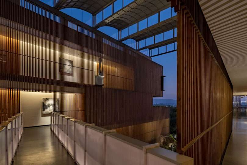 A wide hallway by Malik Architecture features wooden slats, soft lighting, art on the walls, and large windows framing a dusk sky in this modern building.