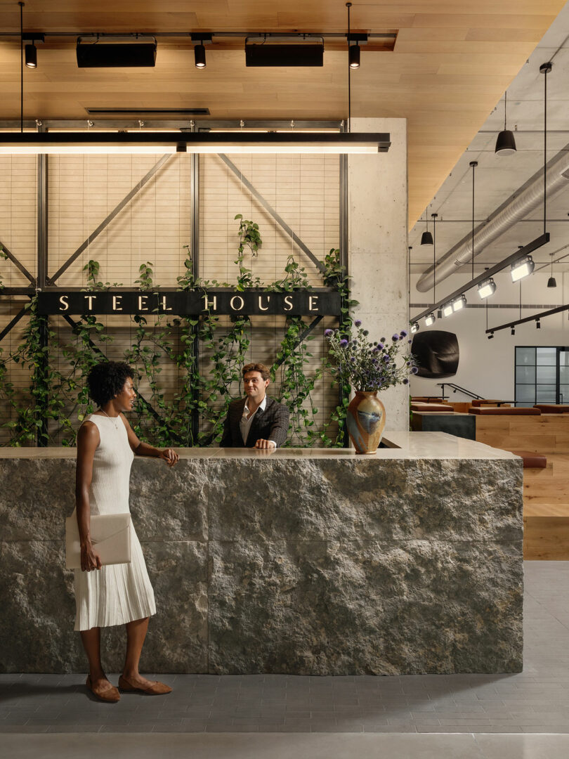 A woman in a white dress talks to a man at a stone reception desk in a modern lobby designed by Morris Adjmi Architects, with 