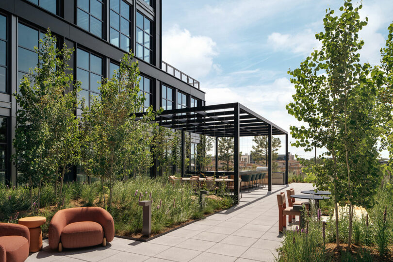 Modern outdoor terrace by Morris Adjmi Architects with seating areas, tables, greenery, and a pergola next to a glass office building under a partly cloudy sky.