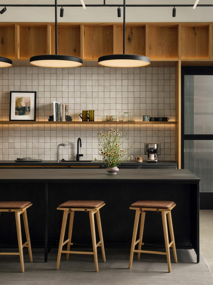 Modern kitchen by Morris Adjmi Architects featuring a black island, three wooden stools, open shelving with decor and glassware, tiled backsplash, and two large pendant lights overhead.